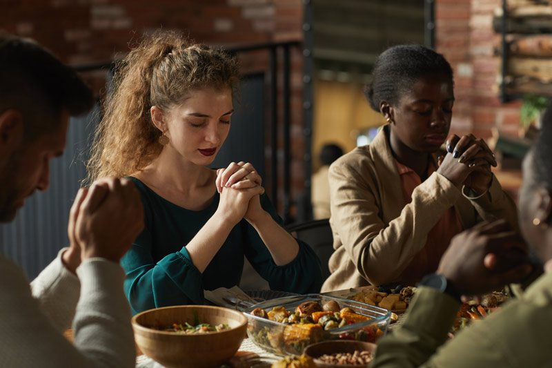 First Holiday Season in Recovery, small, ethnically diverse group of friends seated at a table praying before eating holiday meal - holiday season
