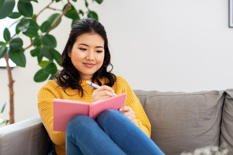 pretty young Asian woman wearing a bright yellow sweater and writing in her pink journal while relaxing on her couch - focus on the process