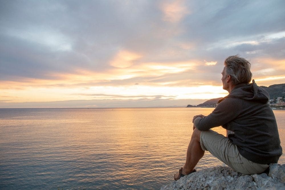 A man sits on a rocky ledge overlooking calm water at sunset, gazing toward the horizon. The sky glows with soft orange and blue tones, creating a peaceful, reflective mood.