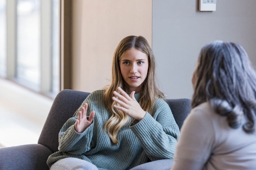A young woman sitting in a chair, gesturing as she speaks with another person during a conversation or counseling session.